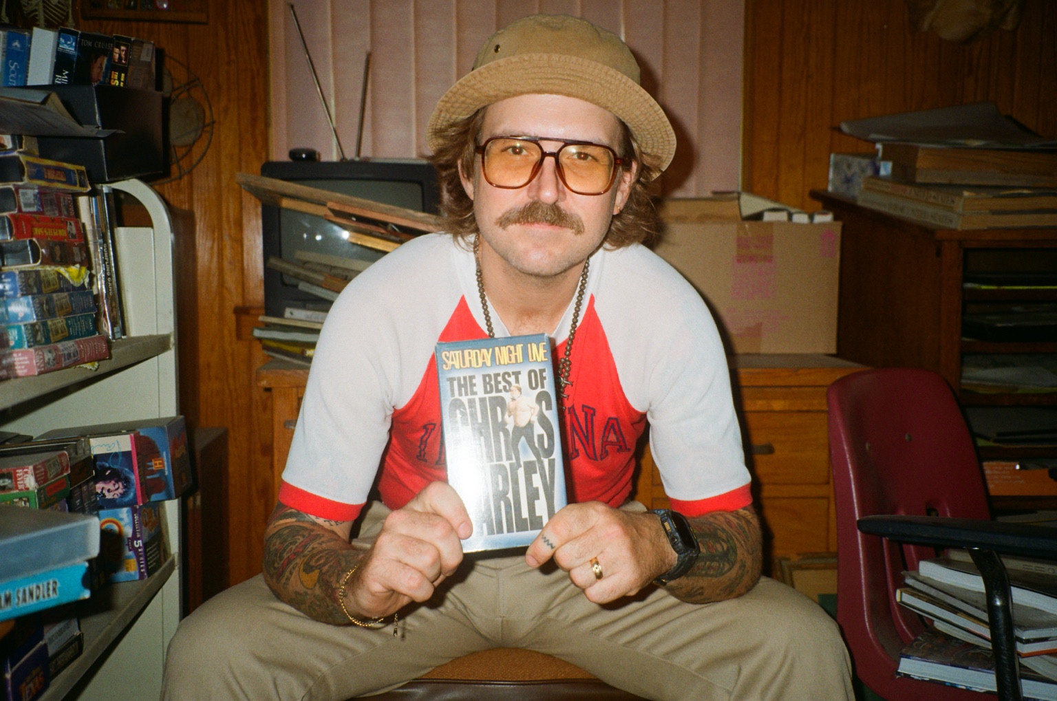 Matt Egan wearing a hat and glasses, holding a Chris Farley VHS tape.